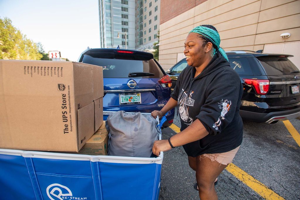 Photo: Ashanti Smith (CAS’24) unloads her car with the help of her mom, not pictured, in order to move into her StuviII apartment. A young black woman with black and teal box braids pushes a large, loaded blue cart away from a parked car.