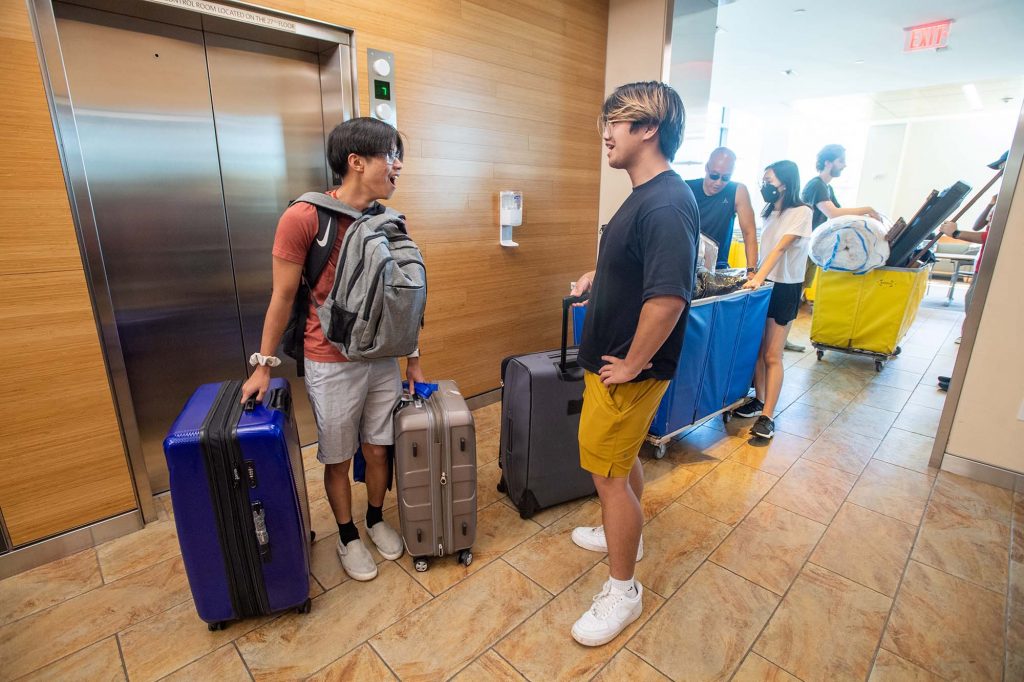 Photo: Ryan Liau (ENG’25), left, and one of his seven roommates Henry Nguyen (ENG’25) wait for an elevator at StuviII in order to move in August 31. Two young Asian men are shown laughing as they roll carryon suitcases in front of an elevator. Relatives are shown in the distance helping to push large yellow and blue bins to the elevator.  Photo by Cydney Scott