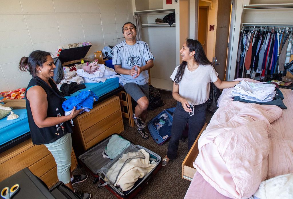 Photo: Shreya Parameshwara (Questrom’26) sets up her Claflin Hall dorm room with the help of her parents; mom, Sandy, and dad, Shivraj. A young Brown woman and her parents laugh and look at each other in her dorm room. The room's spaces are filled with open suitcases and packages as they unpack.