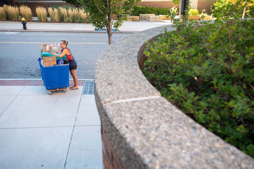 Photo: Christine O’Neill waits for help to bring a packed blue bin to her daughter Nora O’Neill (CAS’26) during move in on BU's West Campus. A white woman wearing a rd shirt and black overalls stands over a large, packed blue bin on an empty sidewalk.