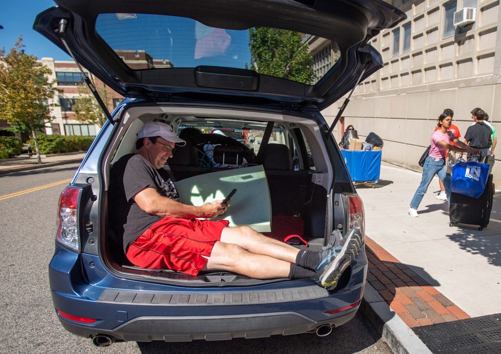 Photo of a BU parent taking a moment to themself as they rest in the open trunk of their mini van and look at their phone.