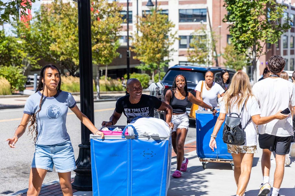 Photo of a Black family helping their child move in to BU's campus. The family uses two large blue carts as they push them down the sidewalk on a sunny day.