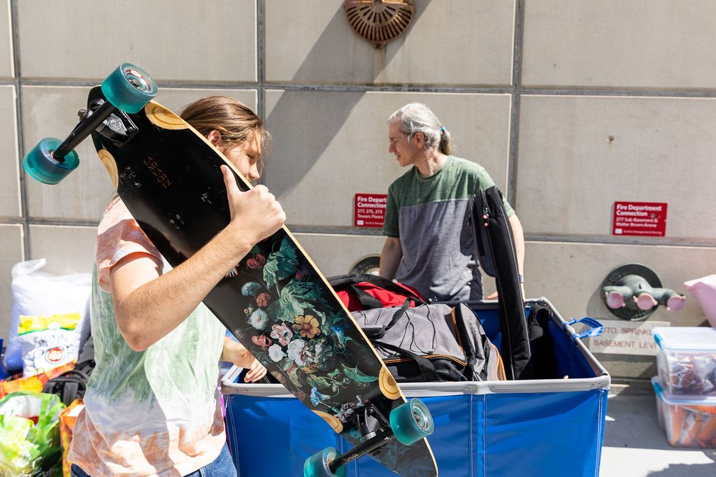 Photo: A student carries an intricately decorated black longboard as they push a filled, large blue cart up the sidewalk.