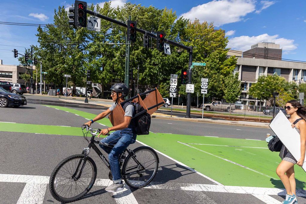 Photo of a BU Student holding a wooden bookcase as they ride their bike down Comm Ave. for move-in. The hold the bookcase over their body as they ride their bike.