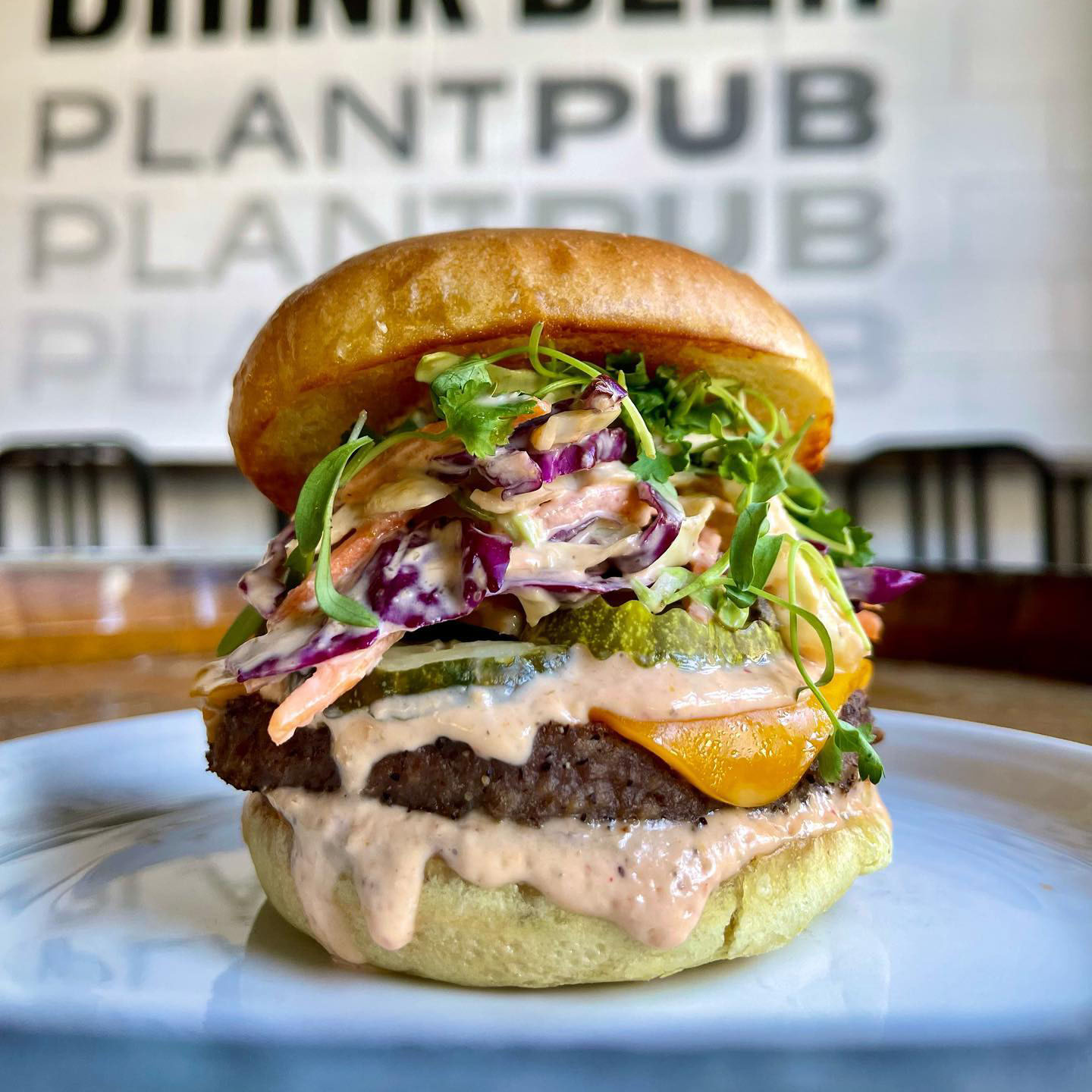 Photo: Closeup of a large plant-based burger with a large patty, slaw, cheese, on a brioche bun. Sign behind it reads "PlantPub"