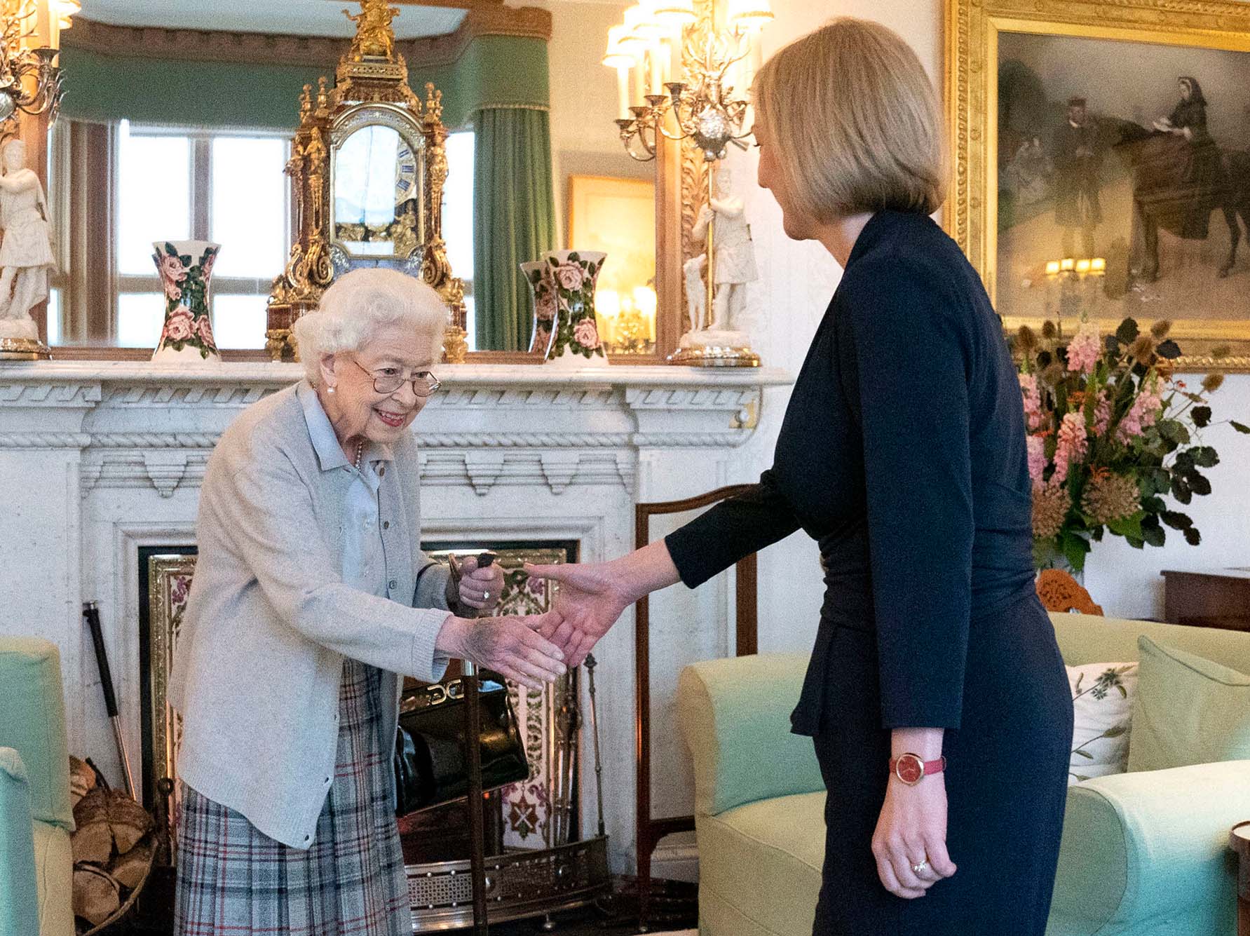 Photo of Britain's Queen Elizabeth II, left, welcoming Liz Truss during an audience at Balmoral, Scotland. A short, elderly woman with white hair wearing a light blue collared shirt, tan blazer, and plaid long skirt (left) shakes hand with a woman with short blonde hair wearing a navy suit (right).