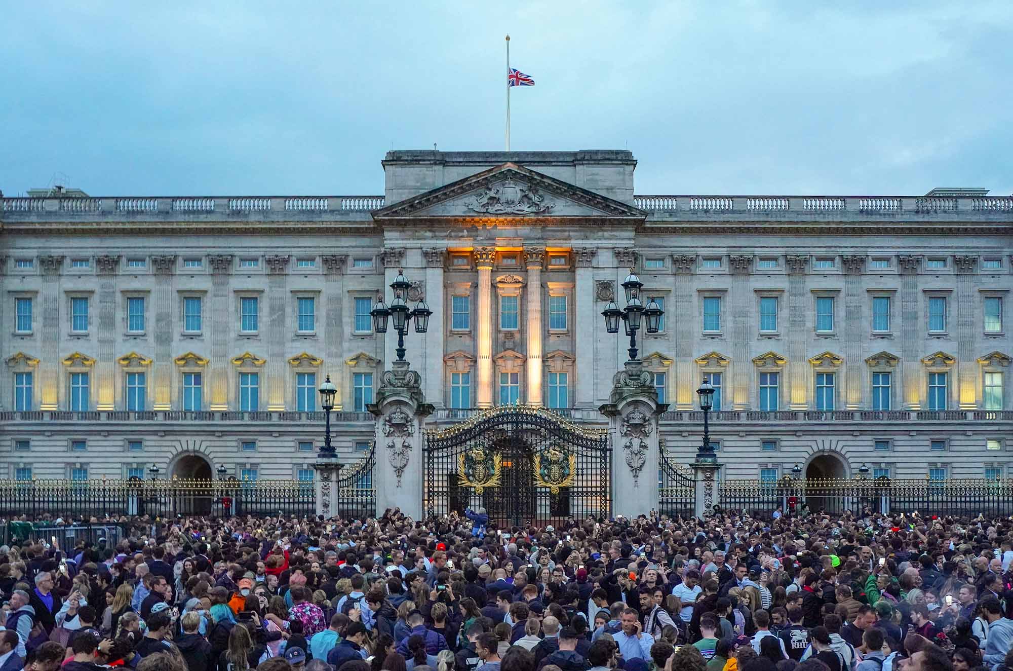 Photo showing a large crowd of people gathered in front of Buckingham Palace during dusk. The UK flags stands atop the palace at half mast, honoring the queen's death.