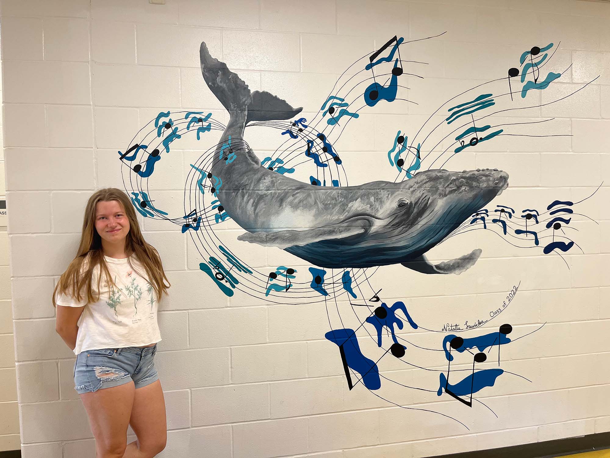 Photo: A young white woman with long blonde hair stands next to a beige wall with a large intricate mural of a whale and music notes flying around said whale.