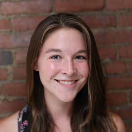 Photo of Teresa Harvey. A young white woman with long, dark brown hair poses and smiles in front of a brick wall.