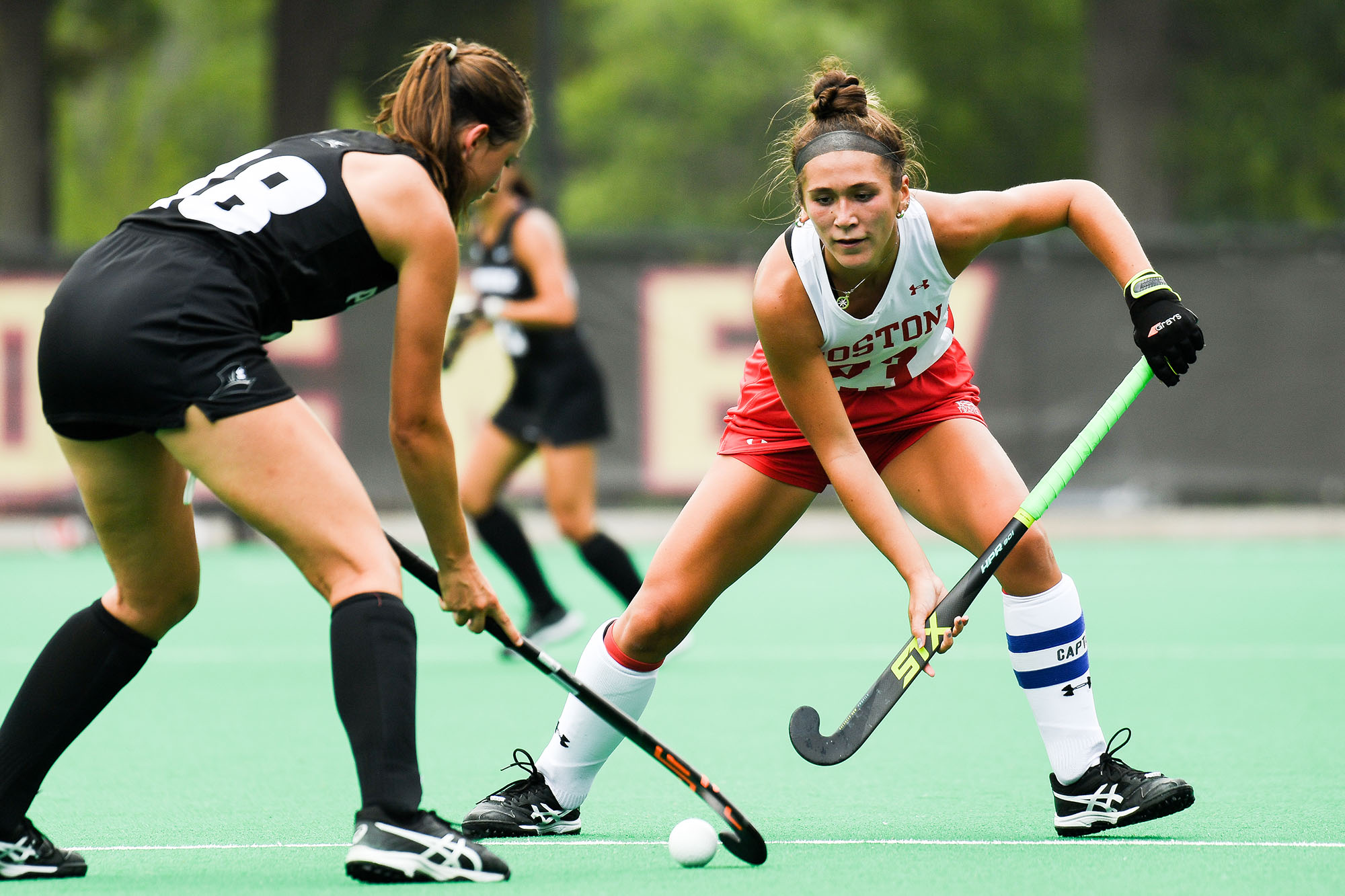 Photo: Midfielder Casey Thompson defends against Providence back Eléonore De Keyser at the Terrier home opener. A young white woman wearing a white and red field hockey uniform is bent over with field hockey stick in hand in front of another white woman in and all black field hockey uniform hitting a small white ball with her field hockey stick.