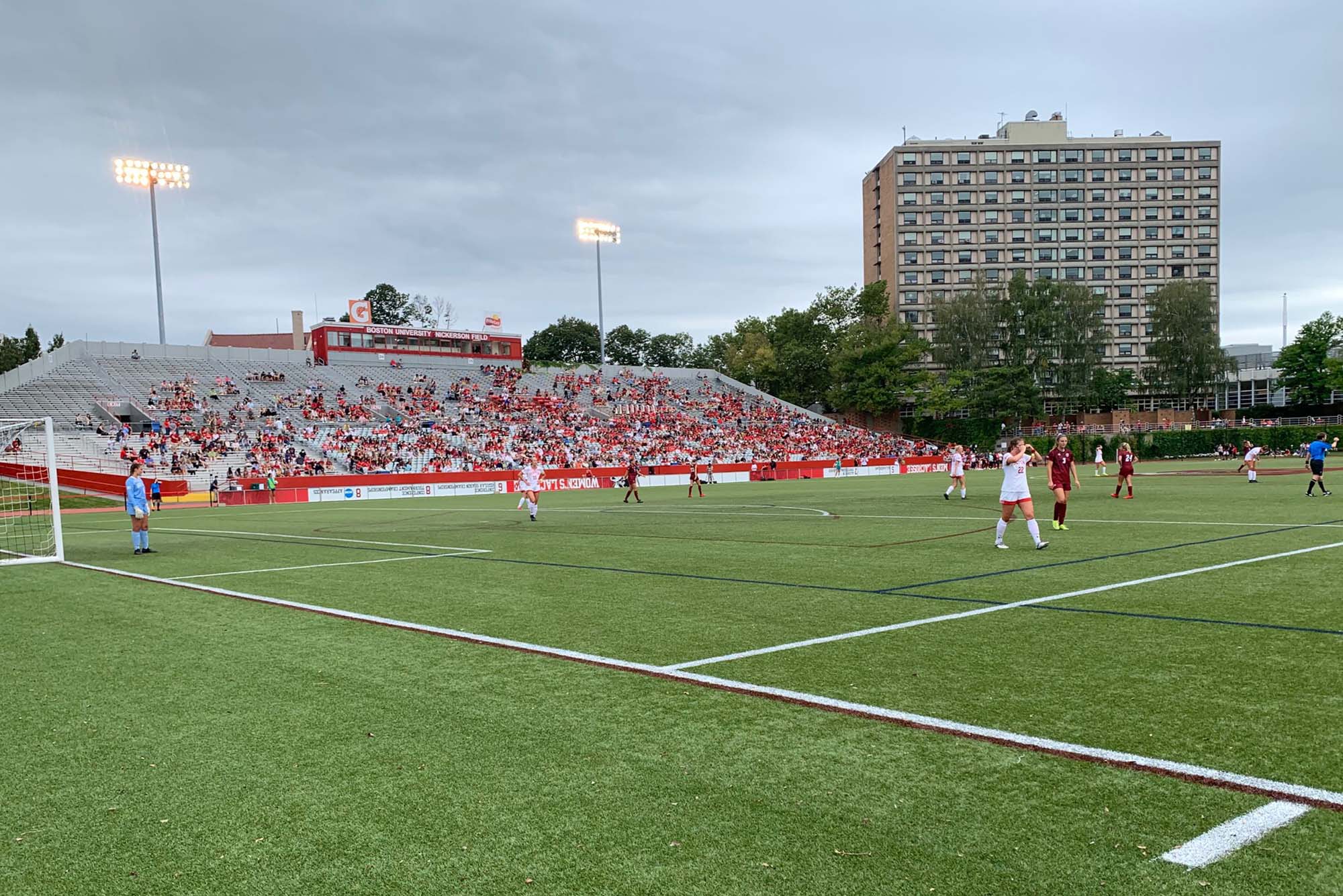 Image of a BU women's soccer match. Players are seen distantly on a field as students wearing red watch int he stands.
