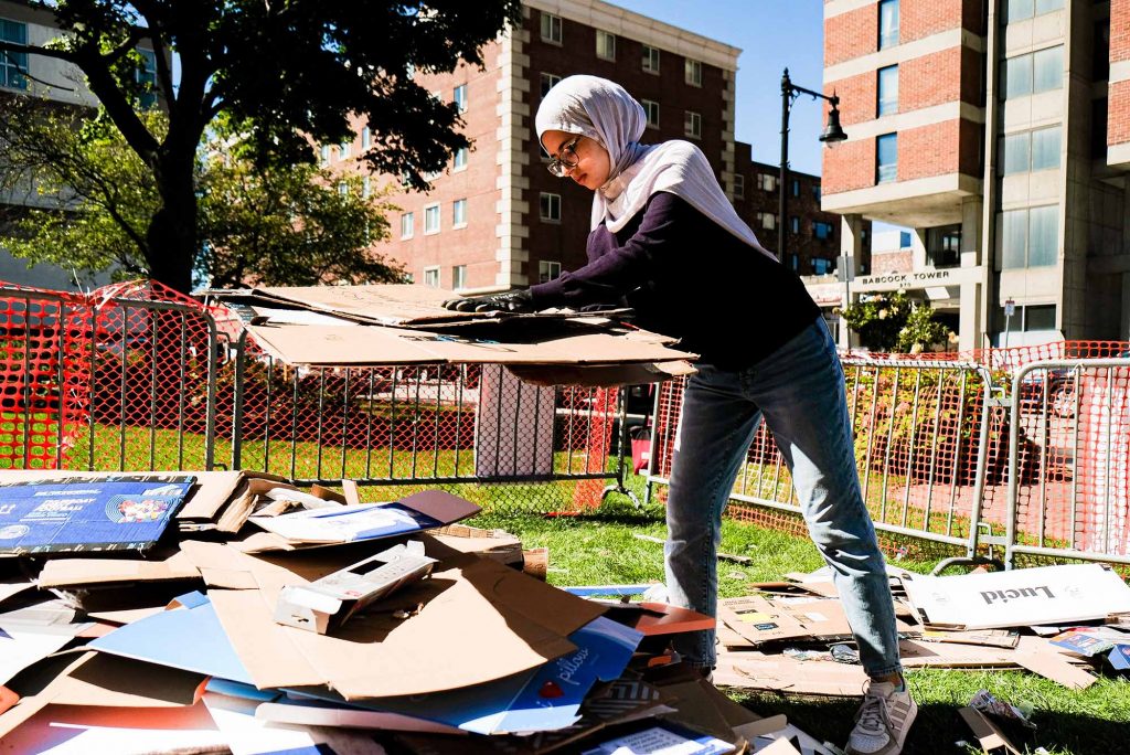 Photo: Ramia Rahmonne (CAS'23) helps collect paperboard for recycle purposes on West Campus. A young woman wearing a hijab helps stacks flattened cardboard boxes on a lawn.