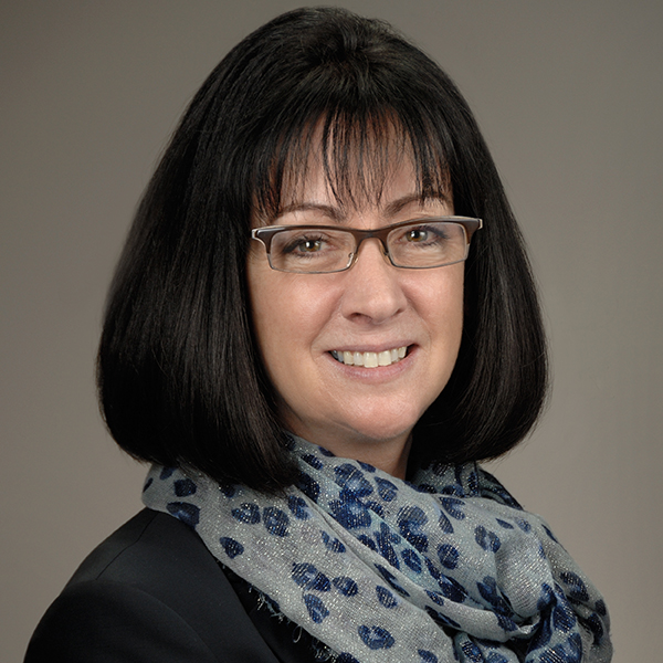 Headshot photo of Nancy J. Sullivan. A white woman with a black bob haircut wearing glasses, a grey and blue floral scarf, and a black top smiles and poses in front of a grey backdrop.