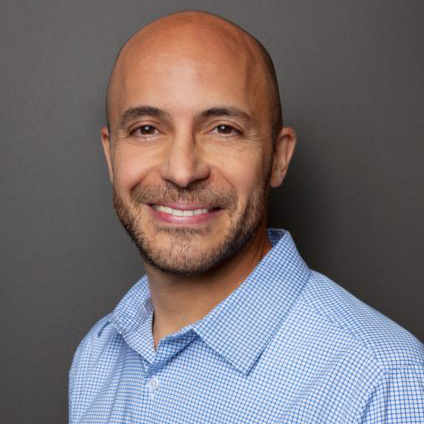 Headshot photo of Professor Michel Anteby. A tan man wearing a light blue collared shirt looks and smiles at the camera in front of a grey backdrop.