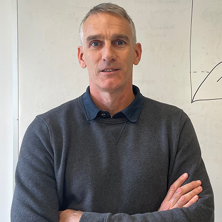 Photo of Peter Blake, a CAS associate professor of psychology. A tall man with grey hair, wearing a navy collared shirt and grey sweater, stands and poses with arms crossed in front of a large whiteboard.
