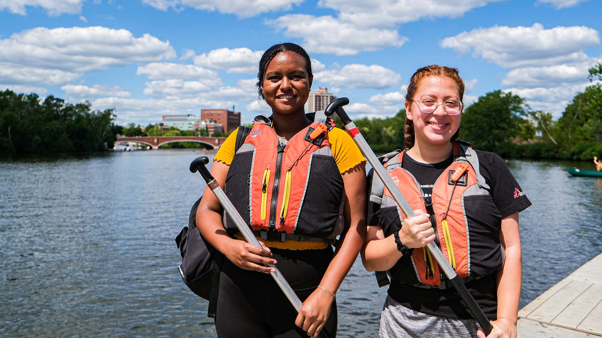 Photo of Isabelle Yap (Sargent'23) , (left), and Genesis Veiassa (Sargent'24) standing on a dock along the Charles River. The Harvard Bridge is seen behind them in the distance; the water and sky are blue with puffy clouds. Yap is a young Black woman with her dark hair pulled back. She has a backpack slung on one shoulder and wears a yellow top and black spandex. Veiassa is a white Woman with long red hair pulled back into french braids, she wears a black t-shirt and gray shorts. Both smile, wear orange, black and gray life jackets, and hold canoeing paddles across their chests.