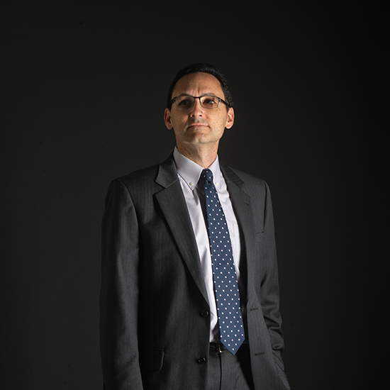 a photo of SPH professor Greg Wellenius. He stands in front of a dark studio background wearing a suit and blue tie with one hand in pocket.