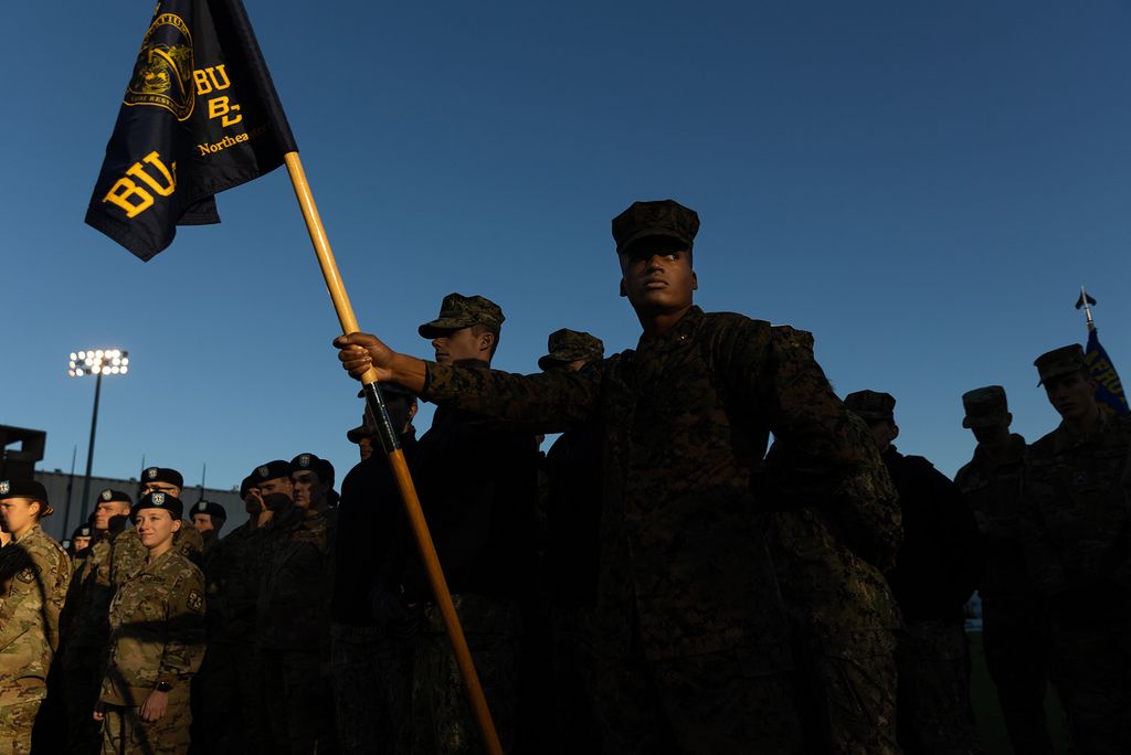 Photo: Marine MIDN First Class, William Reid (CAS'23) listens to Brigadier General Paul L. Minor (not pictured), Assistant Adjutant General (Army) for the Massachusetts National Guard. Students dressed in army camo gear stand at attention. A young Black man stands at front, holding an ROTC flag that reads "BU, BC, Northeastern" and has the ROTC insignia at center. They group of students are shrouded in half shadow, making for an intense morning scene.