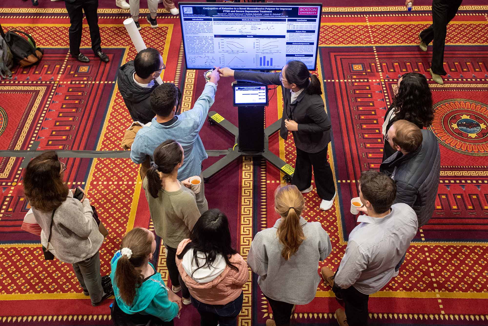 Photo: Siyuan Li (ENG’24), at top in black, presents her work to a small crowd during the 25th Annual Undergraduate Research Symposium in the GSU Metcalf Ballroom Oct 21. Li’s poster was titled "Conjugation of Ketamine to Novel Mucoadhesive Polymer for Improved PTSD and Severe Depression Symptoms".