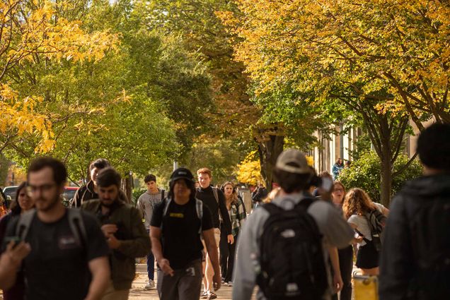 Photo: BU campus stock showing people walking up and down a street as yellow, orange, and red fall leaves line the trees and cover the skyline around them.