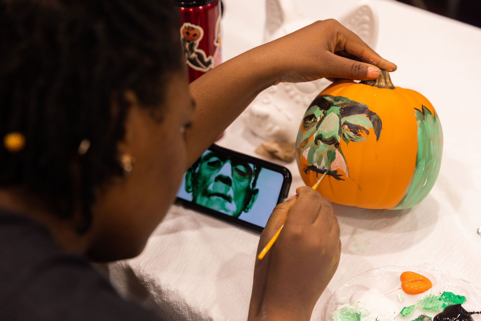 Photo: A young Black person paints Frankenstein's monster onto an orange pumpkin. They use a phone with the picture on the screen (placed in front of them) for reference.