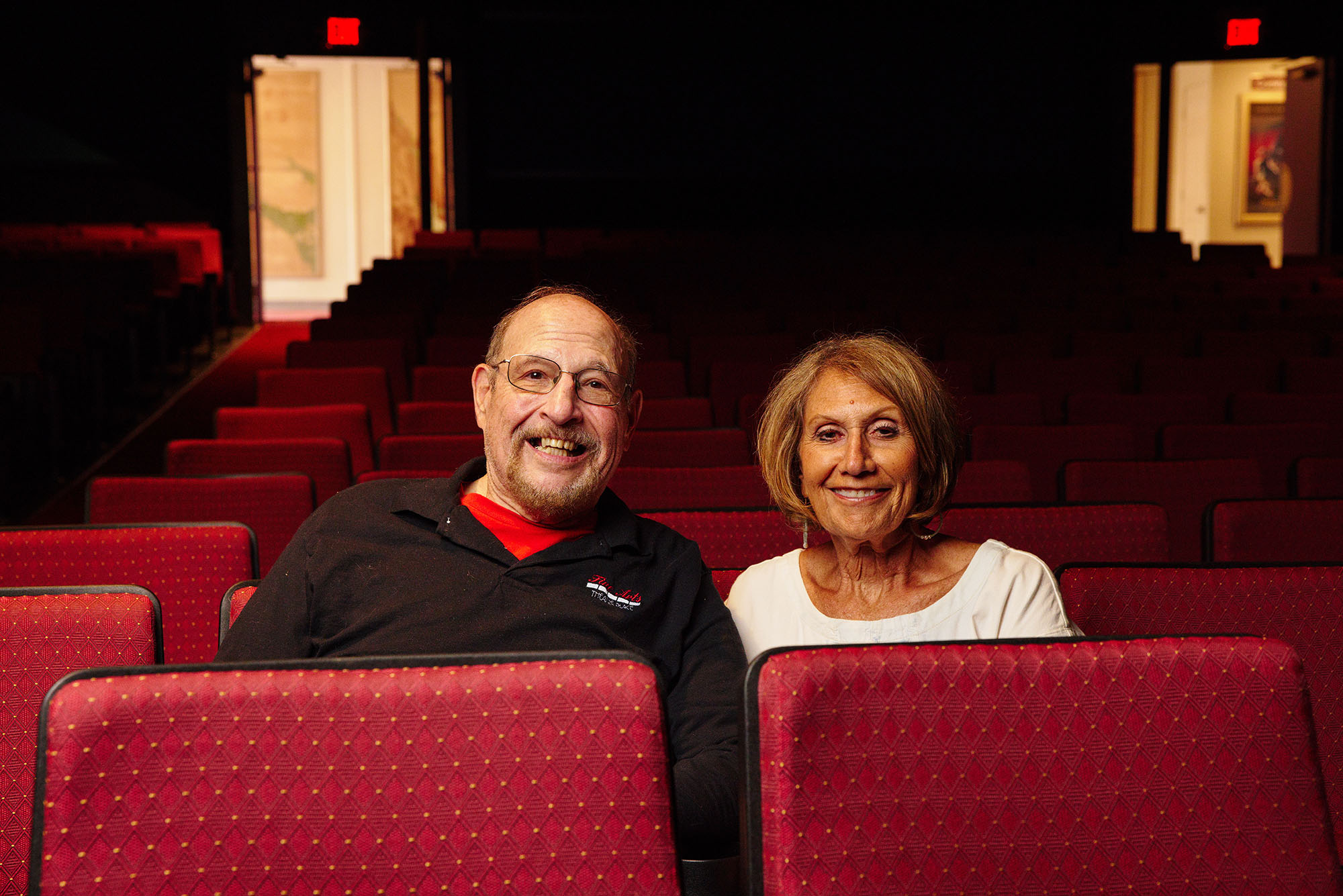 Photo: Alums Les Gordon (left) and Dafna Krouk-Gordon sit in an empty theater and pose for the camera. An older white man wearing glasses and a black long sleeved shirt sits tot he left of an older white woman wearing a white top. They both sit in an empty theater with red seats and smile for the camera.