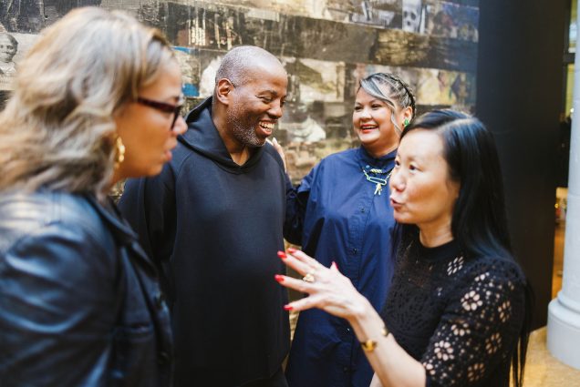 Photo of Cey Adams (center, left) and show curator Liza Quiñonez (center, right) at the opening reception for the first-ever retrospective exhibition of Adams’ work, Cey Adams, Departure: 40 Years of Art and Design at the Faye G., Jo, and James Stone Gallery, September 30. In the foreground: Makeeba McCreary (left) and Linda Yang. Four people talk and laugh in an art gallery.