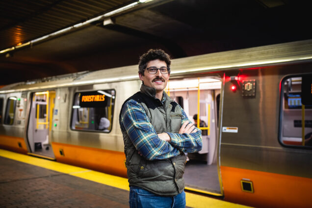 Photo of Josh Fairchild standing in front of an Orange line train with its doors open for boarding. a white man wearing a blue plaid shirt and grey puffer vest poses with arms crossed in front of an Orange and silver train.
