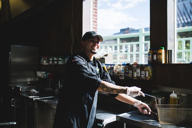 Photo: Marcial Matos, a Puerto Rican cook at Granby kitchen in the Hillel House, smiles as he grabs hold of a fryer basket in a dark kitchen. A large window lets in light behind him.