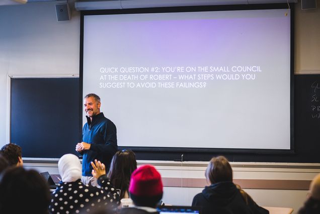 Photo: Professor Phillip Haberkern teaches a CAS history class "Game of Thrones: Power and Politics in Pre-Modern Europe “ on October 19, 2022. A white man with a grey beard and head of hair stands in front of a classroom and smiles as students look on. Behind him, the projection on screen reads "Quick question #2: You're on the small council at the death of Robert - what steps would you suggest to avoid these failings?"