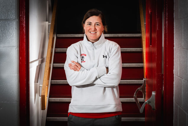 Photo: Posed photo of Casey Brown, BU women's soccer head coach. Brown stands and smiles with arms crossed in an open doorway before a set of stairs behind her. She wears a white long sleeved shirt with the BU soccer logo on it.