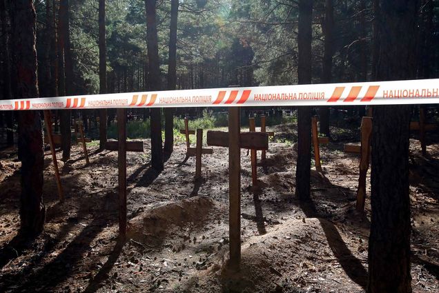 Photo: Wooden crosses mark the graves of Izium residents killed by Russian occupiers during exhumation procedures at a mass burial site in Izium that was liberated by the Ukrainian Armed Forces from Russian troops, Kharkiv Region, northeastern Ukraine. Numerous mounds of dirt are marked by crosses in a wooded area. A tape is pulled across the scene, with Ukrainian writing on it.