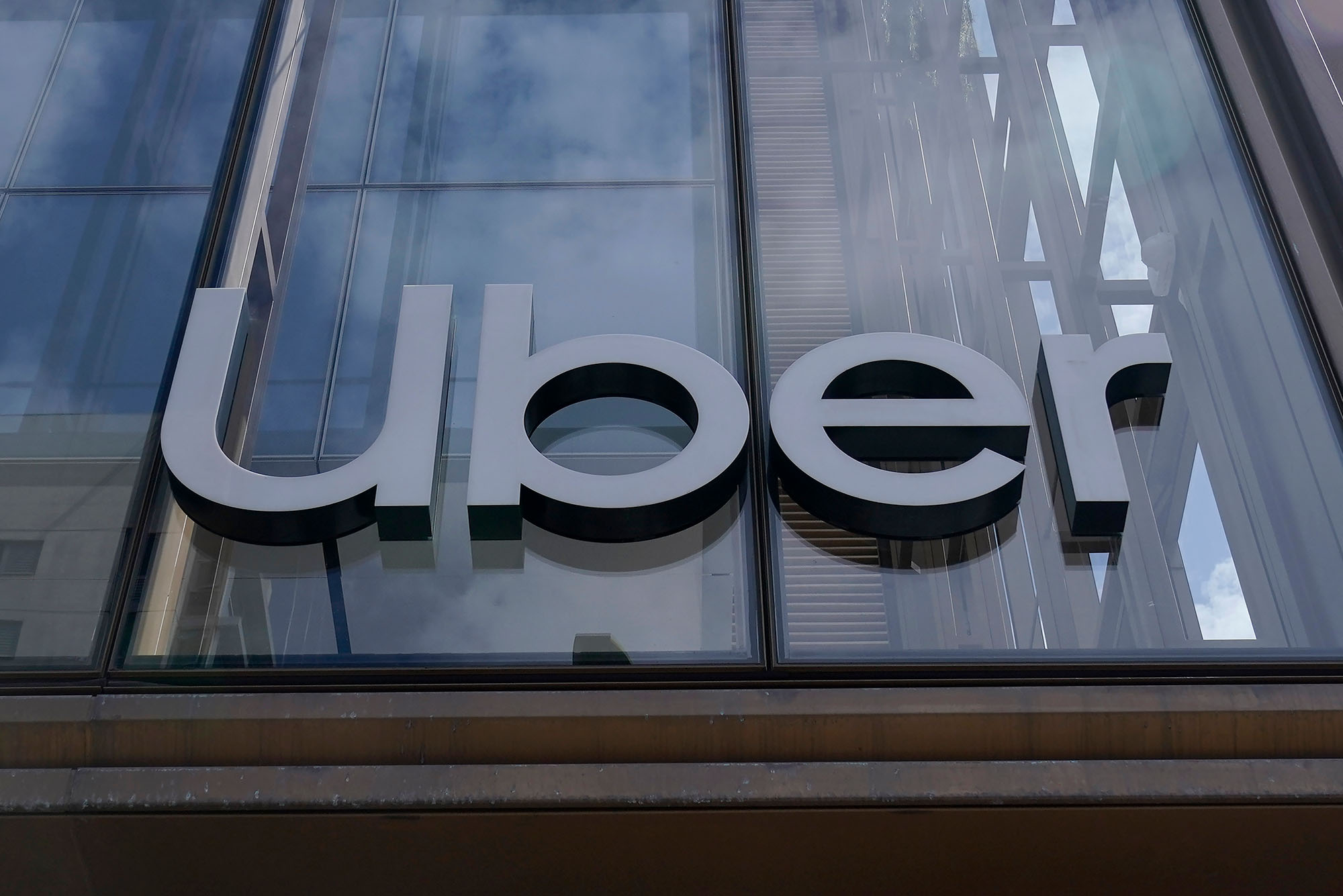 Photo of an Uber sign is displayed at the company's headquarters in San Francisco. Metal sign reads "Uber" on a glass-paned building.