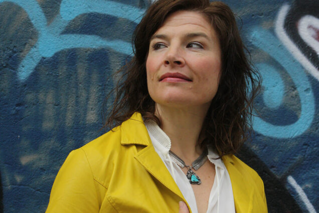 Photo: Jenn Lindsay poses for a photo. A white woman with wavy brown hair and wearing a white collared shirt and yellow jacket poses in front of a blue wall and looks to the left.