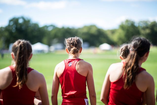 Photo: The backs of the three women BU cross country runners are shown as they line up at the start of a race. They face a vast green field and wear BU track and field jerseys.