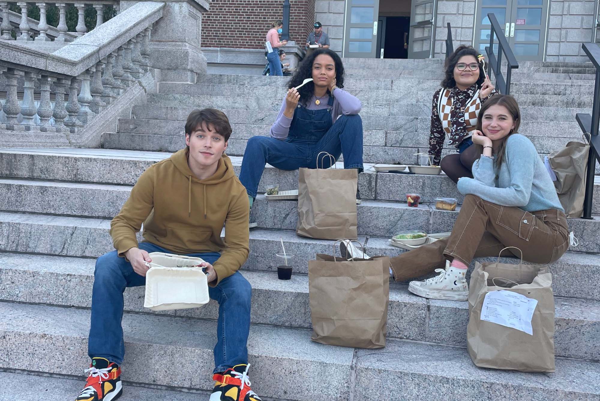 Photo of 4 young adults sitting on a large set of stone steps. They all smile and strike a quick pose for the camera on a cloudy day. Large brown paper bags sit at each of their feet.