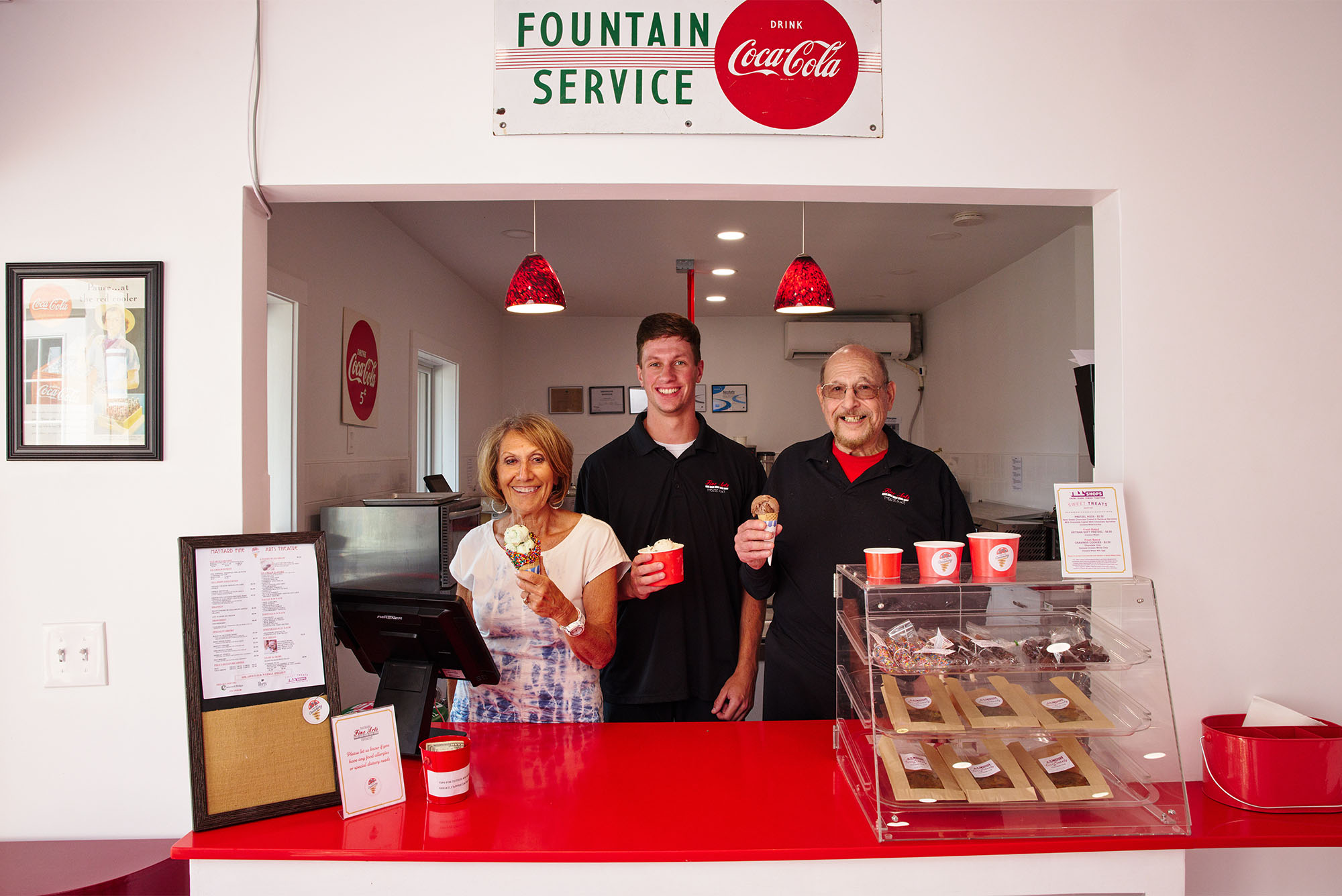 Photo of a Creamery inside the theater. Les Gordon and Dafna Krouk-Gordon flank Joey Livolsi. Three people stand behind a red counter holding paper bowls of ice cream as the smile and pose. The sign above them reads "Fountain Service" and features a large coca-cola logo.