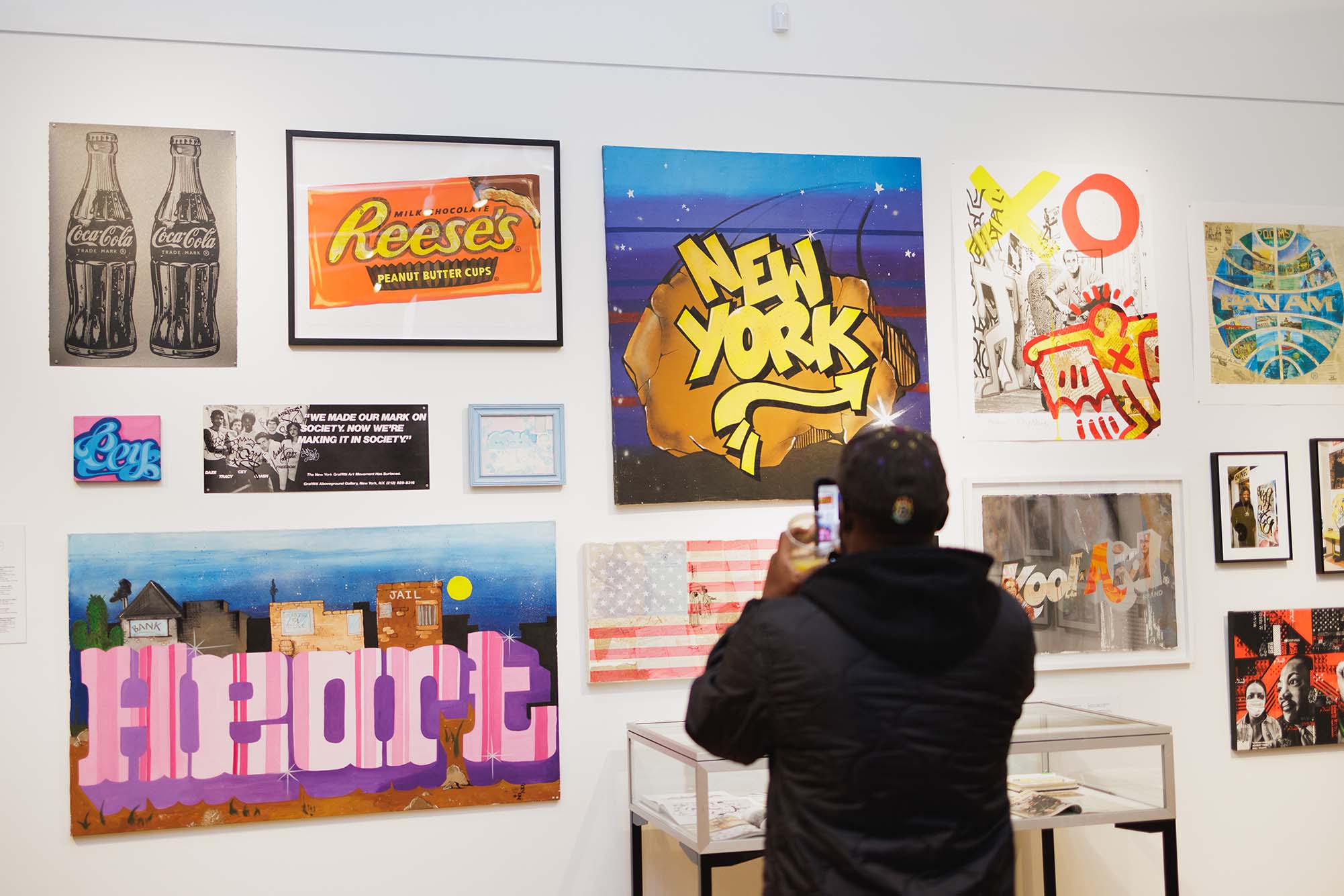 Photo: A person snaps a photo on their phone of a gallery wall in Cey Adams' exhibit. Wall shows various graffiti works and depictions.