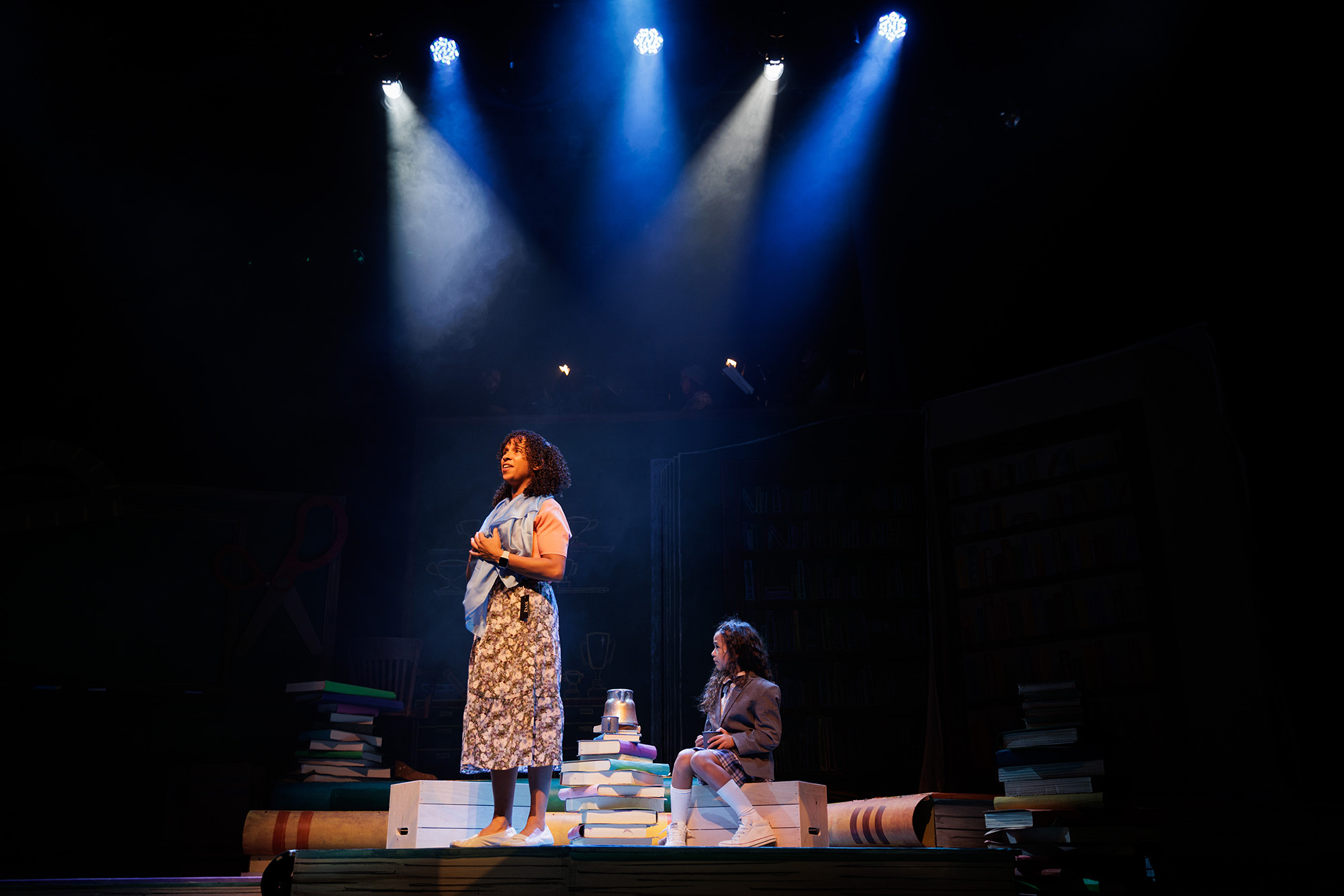 Photo: Kira Troilo (left) and Sky Fuller share a quiet moment on stage. A young woman with curly hair and wearing a simple, floral dress stands and looks to the left on stage as a young girl wearing a brown school uniform sits on a crate to her right and looks on. The stage is light by only blue lights, relaying a somber mood.