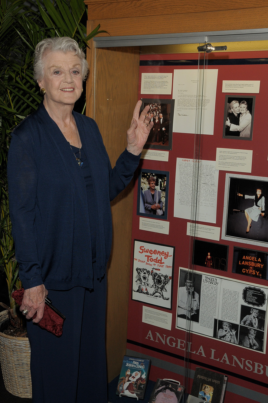 Photo:  Lansbury in 2011 in front of a case of memorabilia from her career on view at the Howard Gotlieb Archival Research Center. An older white woman with graying hair poses and points to a large glass case featuring various works.