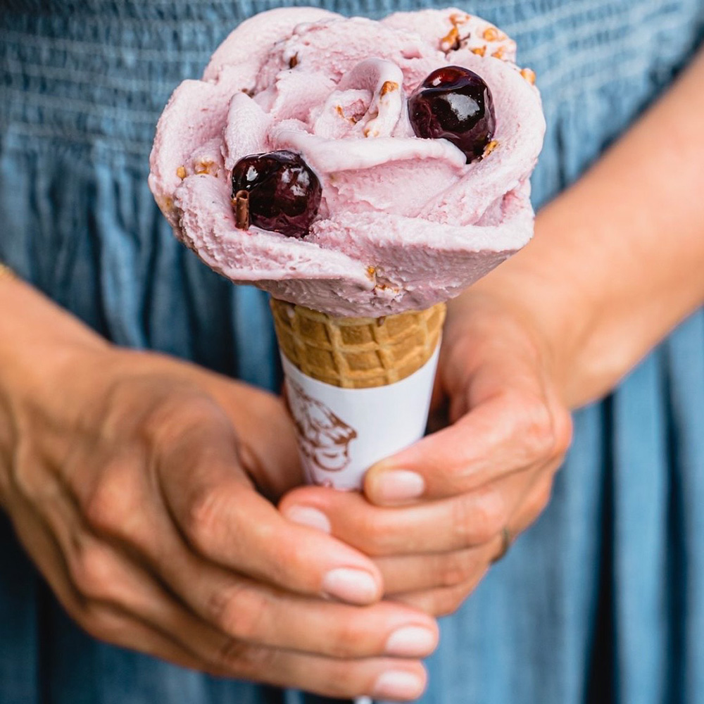 Photo of ice cream cone. Pale purple ice cream is scopped onto a waffle cone in the shape of a rose and topped with glazed blueberries.