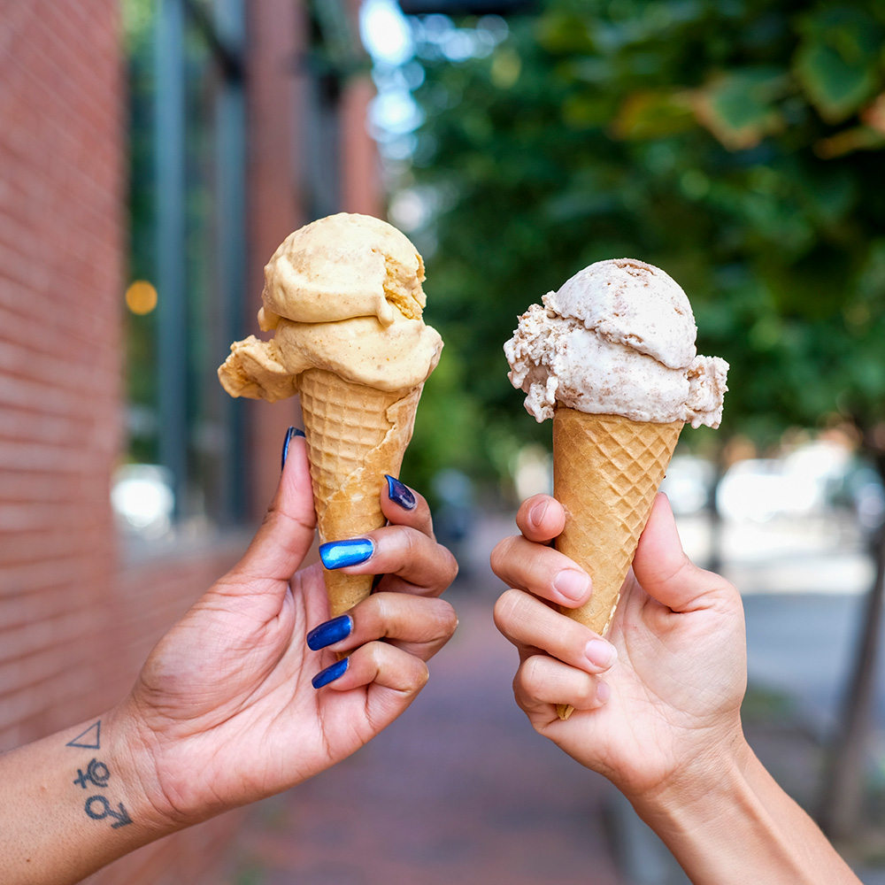 Photo of two hands holding up to waffle cones with scoops of ice cream.