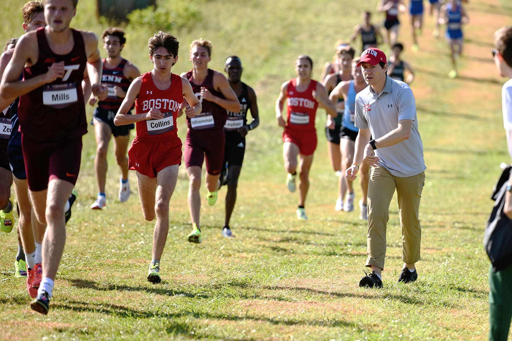 Photo: A group of young men race towards the camera on a grassy field. The lead person wears the scarlet BU cross country jersey and shorts as other wearing Navy uniforms and similar BU uniforms race after him. On the right, a man wearing khaki pants, a red BY cap, and light gray collared shirt, cheers on the side.