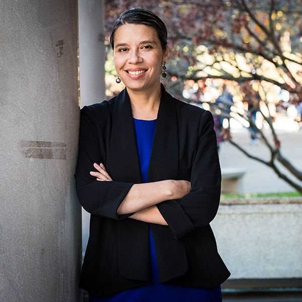 Photo: Jasmine Gonzales Rose poses against a pillar. A Latina woman poses with arms crossed and wears a black blazer and navy blue blouse.