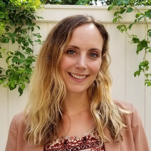 Photo of Alyssa Harlow. A young white woman with wavy, blonde hair smiles in front of a white fence with greenery on its edges.