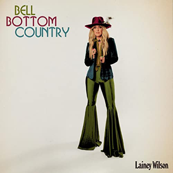 Album Cover shows Lainey Wilson standing and posing in a beige room. She wears olive green bell bottoms pants, a red, large brimmed velvet hat, and a navy jacket. Text reads "Bell Bottom Country".