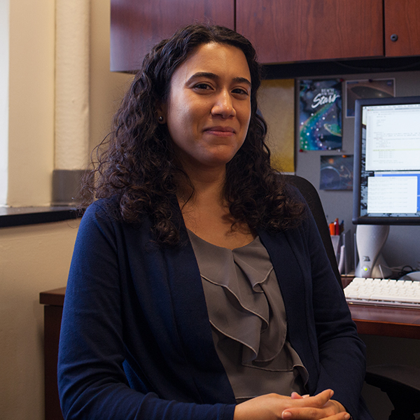 Photo of Catherine Espaillat. A Latina woman with curly black hair poses in a chair at her desk. She wears a dark grey blouse and navy sweater.