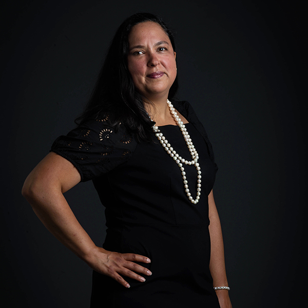 Photo of Diana M. Ceballos. A Latina woman wearing a black dress and a long pearl necklace poses with one hand on hip in front of a dark grey backdrop.