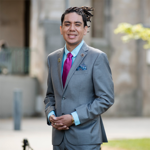 Photo of Daniel Jacobson López. A Latinx person wearing a grey suit ensemble with dreadlocked hair pulled back smiles and poses with hands clasped in front of him on a scenic lawn.