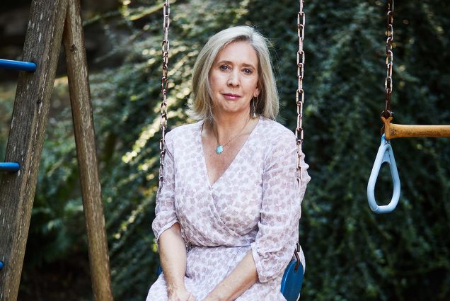 Photo of Warren Binford, wearing a white and pink dress with a spotted pattern, sitting on a kid's swing set outside in someone's back yard. She smiles slightly, a bit somber, and has blonde shoulder-length hair.